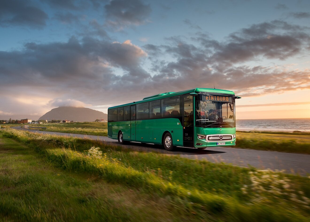 An electric Mercedes-Benz eIntouro inter-city bus drives through a coastal landscape at sunset.