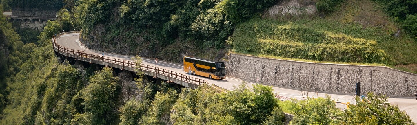 A Setra touring coach drives on a mountain road through a wooded landscape.