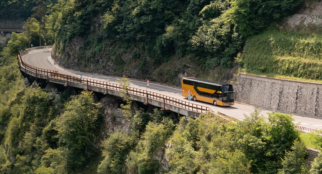 A yellow Setra touring coach drives on a winding mountain road through a wooded landscape.